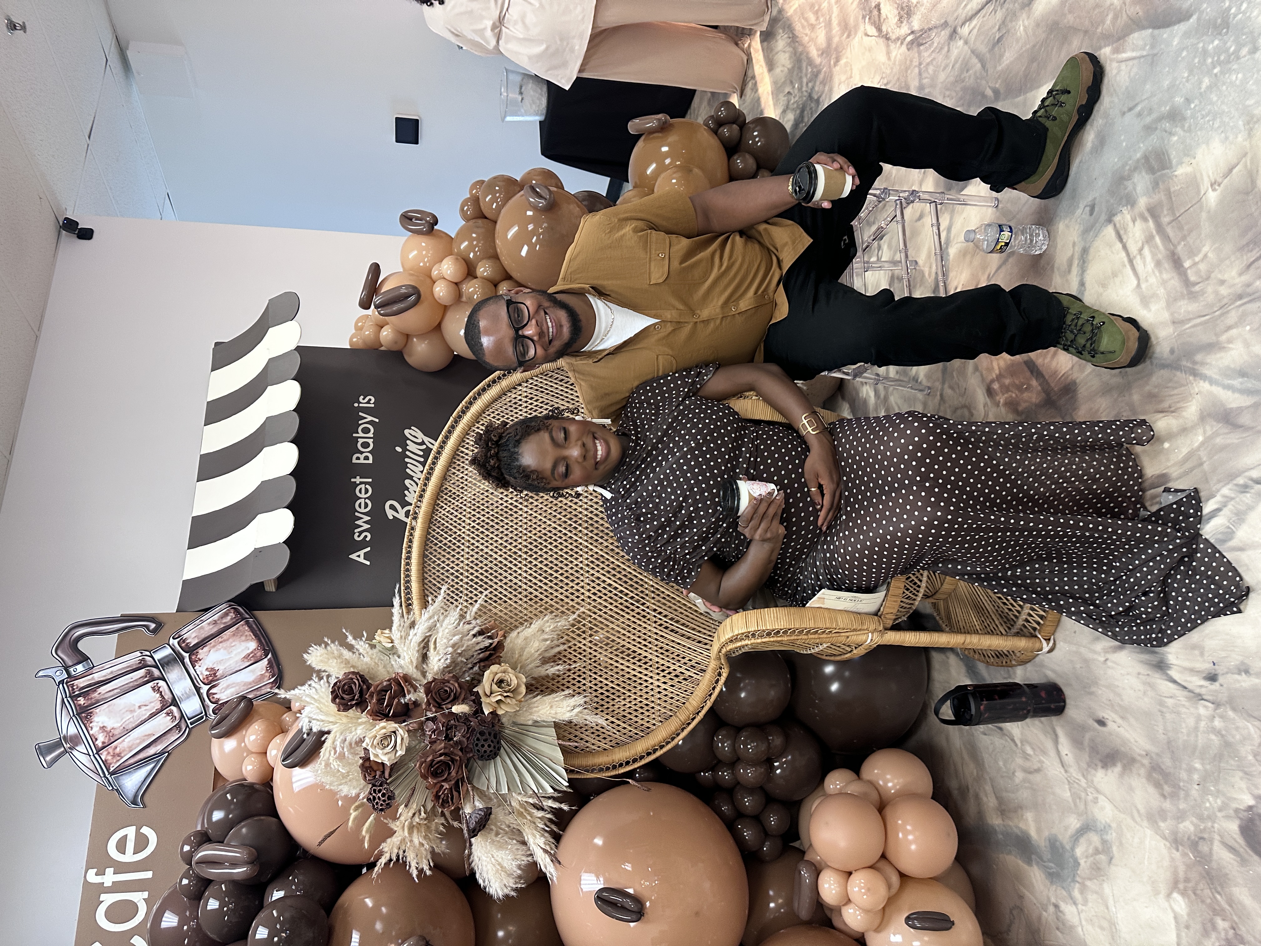 Expectant parents holding Tee Coffee Studio branded lattes at coffee-themed baby shower in Atlanta with balloon arch backdrop reading A Sweet Baby is Brewing